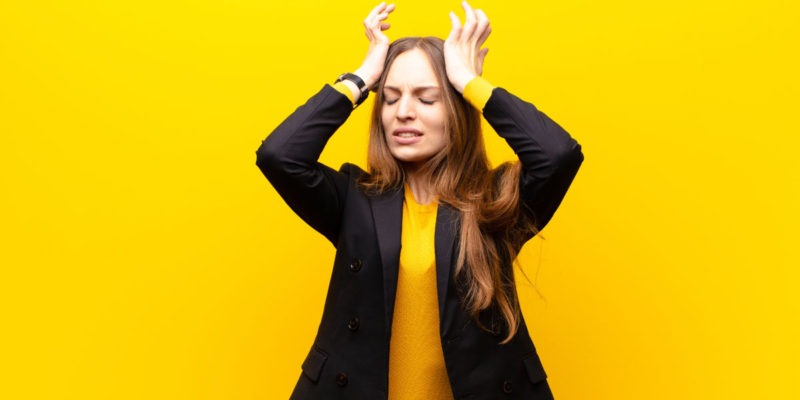 Foto de uma mulher em fundo amarelo com as duas mãos na cabeça demonstrando ansiedade.