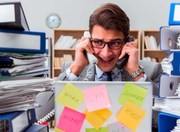 foto de homem no trabalho atendendo dois telefones e com pilhas de relatórios ao lado. seu semblante é de desespero