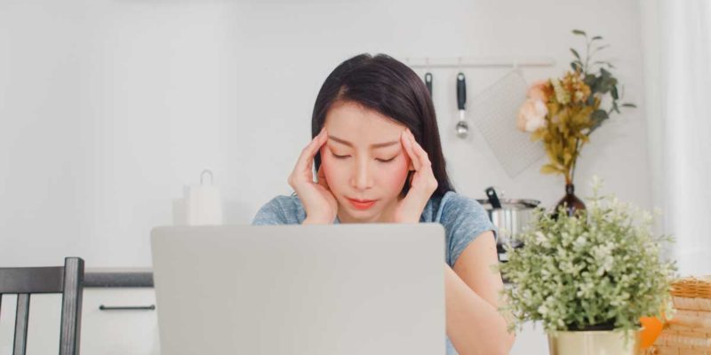 Foto de uma mulher em frente ao computador com as mãos na cabeça e semblante estressado e cansado