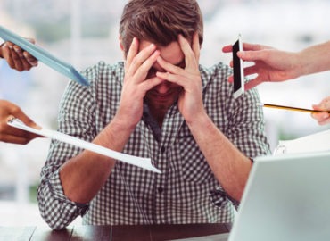 Foto de um homem em sua mesa de trabalho com as mãos na cabeça, olhando para baixo. Várias mãos estão apontando para ele com solicitações de trabalho.