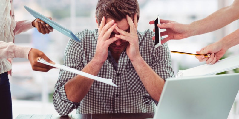 Foto de um homem em sua mesa de trabalho com as mãos na cabeça, olhando para baixo. Várias mãos estão apontando para ele com solicitações de trabalho.