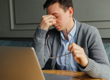 Foto de um homem em sua mesa de trabalho, com um computador à frente, uma das mãos encosta no nariz e os olhos estão fechados. Seu semblante é cansado.