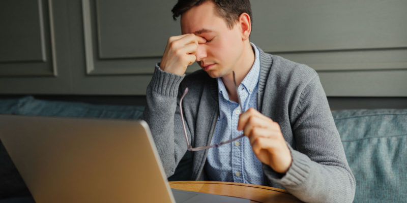 Foto de um homem em sua mesa de trabalho, com um computador à frente, uma das mãos encosta no nariz e os olhos estão fechados. Seu semblante é cansado.