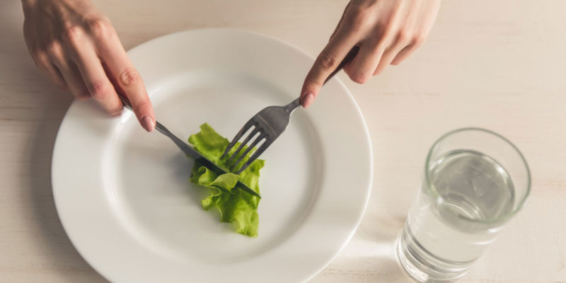 foto de um prato de comida, com uma folha de alface dentro, com garfo e faca prestes a cortá-la