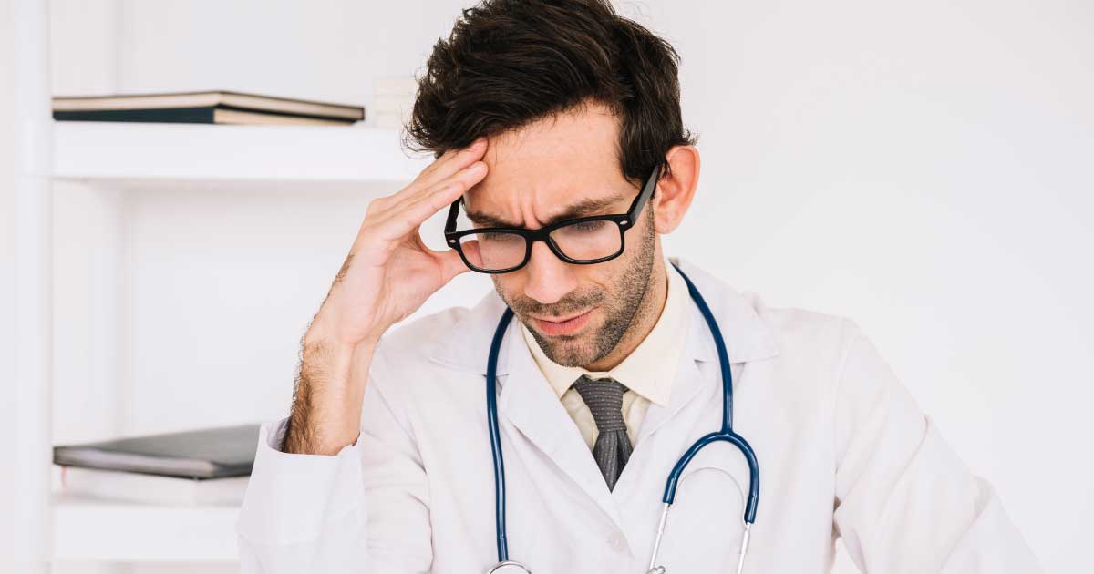 foto de um médico em seu trabalho com a mão direita na cabeça e semblante estressado e preocupado