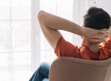 Foto de um homem de costas sentado em uma cadeira olhando para a janela. Ele está com as mãos na nuca em posição relaxada.