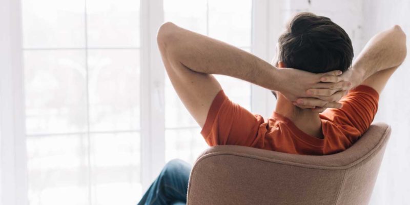 Foto de um homem de costas sentado em uma cadeira olhando para a janela. Ele está com as mãos na nuca em posição relaxada.