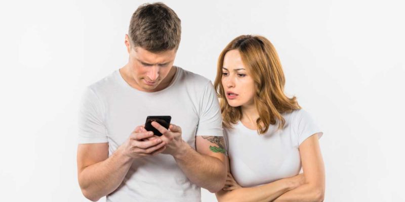 foto de um homem e uma mulher, lado a lado, o homem está mexendo em seu celular e a mulher está tentando ver o que ele está fazendo, demonstrando ciúme e curiosidade