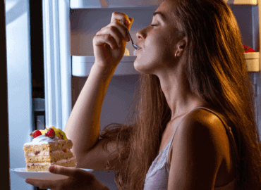 Foto de uma mulher com a geladeira aberta, á noite, comendo um pedaço de bolo de forma compulsiva