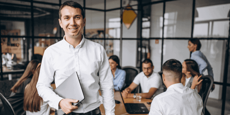 foto de um homem em pé sorrindo e com uma pasta na mão. Atrás dele estão vários colegas de trabalho sentados em uma mesa de reunião