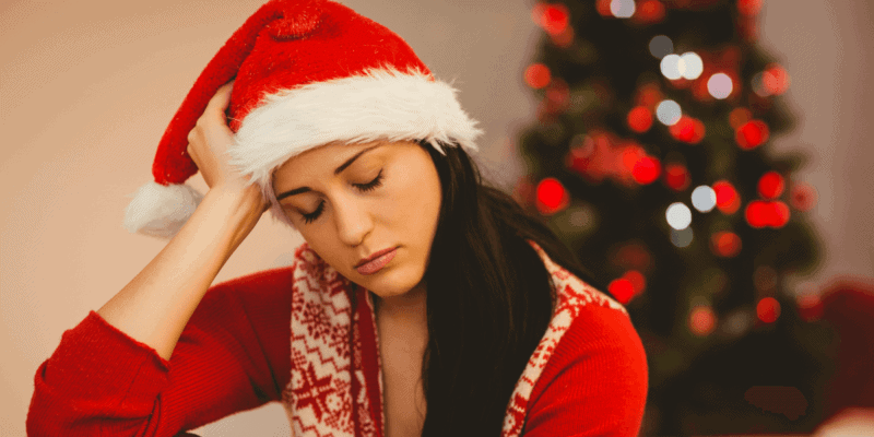 foto de uma mulher sentada na sala, com uma das mãos na cabeça e olhando para baixo com semblante triste. atrás dela tem uma árvore de Natal.